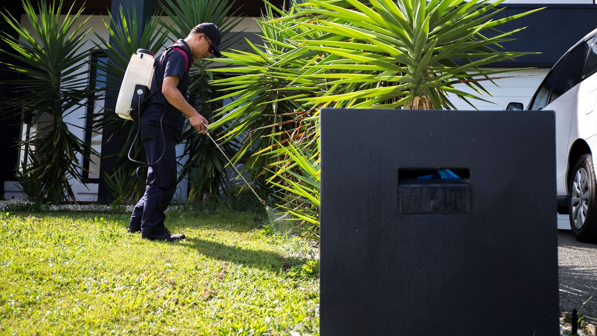 Pest control worker spraying plants near a house and car